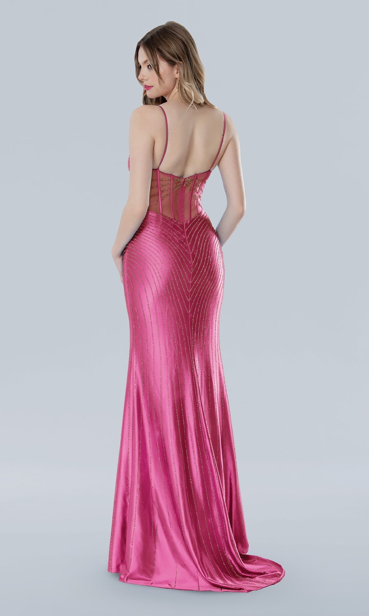 A woman models the Stella Couture 25116, a shiny, floor-length hot pink prom dress with thin straps and a sheer back panel, standing against a plain light gray background.